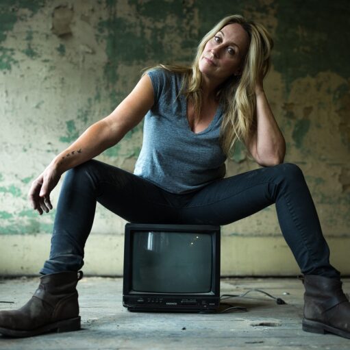 An image of singer-songwriter Amy Speace sitting on top of an old black television set. She is wearing a light blue-grey v-neck t-shirt and dark jeans. The background is a worn-out looking room with peeling paint on the walls.