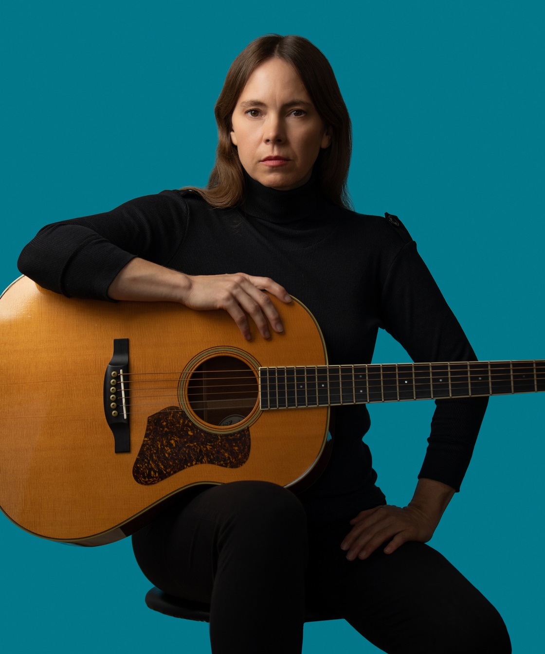 An image of singer-songwriter Bronwynne Brent sitting on a stool and holding an acoustic guitar. She's wearing a black long-sleeved turtleneck and black trousers and is looking directly at the camera. The background is solid turquoise.