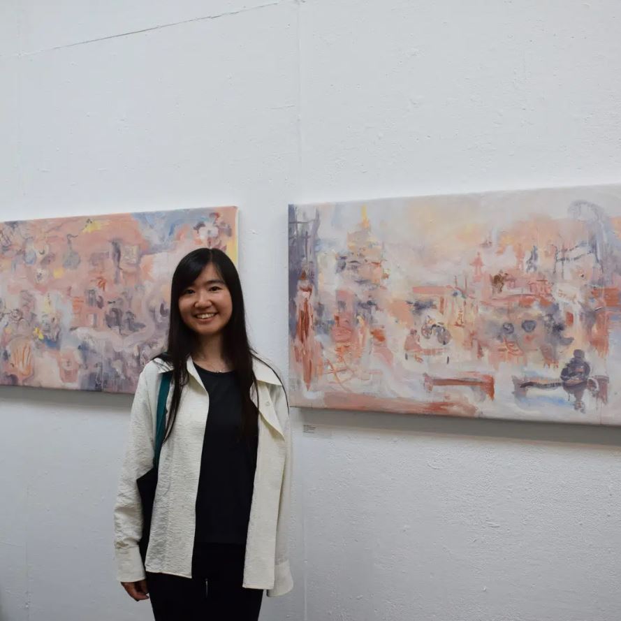 A young woman with long, dark hair stands in an art gallery, smiling at the camera. She is wearing a black shirt and black pants with a white jacket over top. Behind her on the white wall are two horizontal paintings with abstract designs in muted colors like pink, orange, blue, and purple. The paintings are similar in style and are hanging side-by-side.
