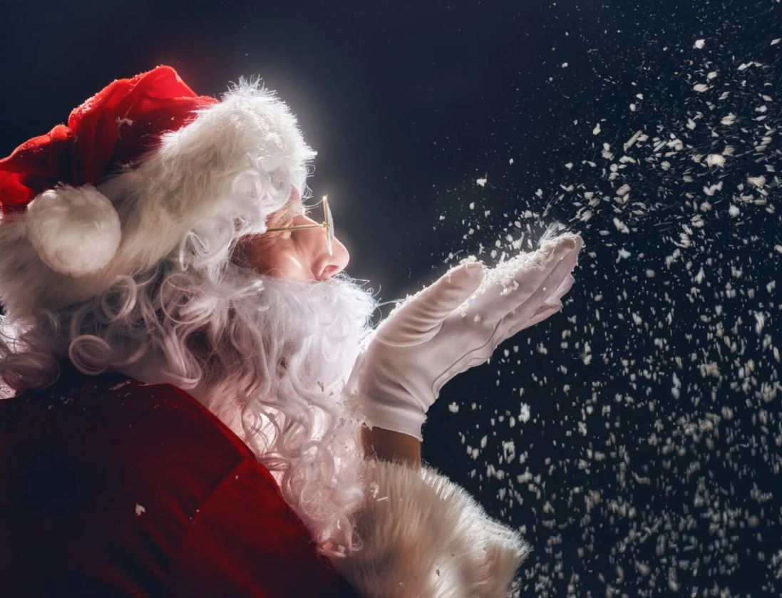A person dressed as Santa Claus is shown in profile against a dark background, blowing a handful of white flakes into the air. The flakes look like snow or glitter and are sparkling in the light. Santa is wearing his classic red and white suit, a red hat with a white pompom, a white beard, and white gloves. He is also wearing small, round glasses.