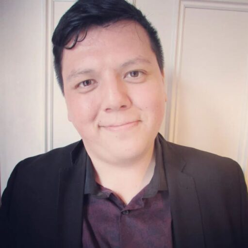 A headshot of a person with short, dark hair, wearing a black blazer and a dark patterned shirt. They are smiling slightly and looking directly at the camera. They are in front of a white wall with a door.