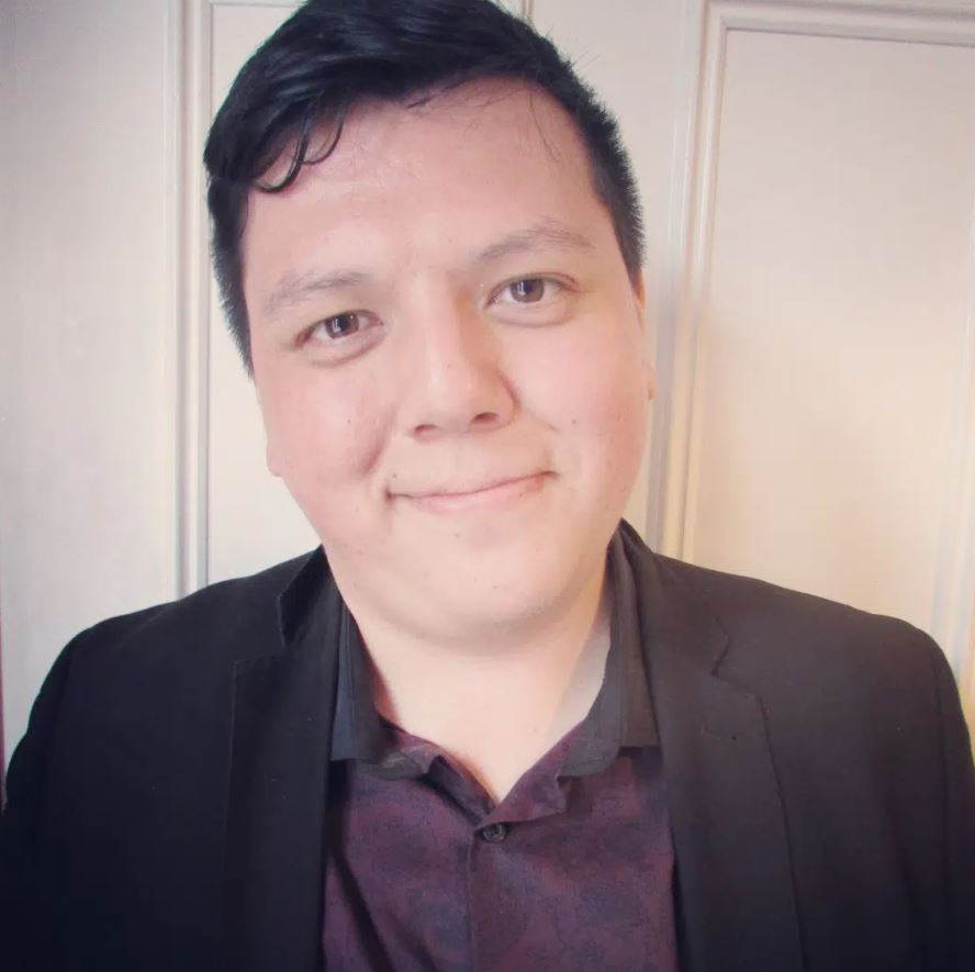 A headshot of a person with short, dark hair, wearing a black blazer and a dark patterned shirt. They are smiling slightly and looking directly at the camera. They are in front of a white wall with a door.