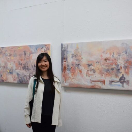 A young woman with long, dark hair stands in an art gallery, smiling at the camera. She is wearing a black shirt and black pants with a white jacket over top. Behind her on the white wall are two horizontal paintings with abstract designs in muted colors like pink, orange, blue, and purple. The paintings are similar in style and are hanging side-by-side.