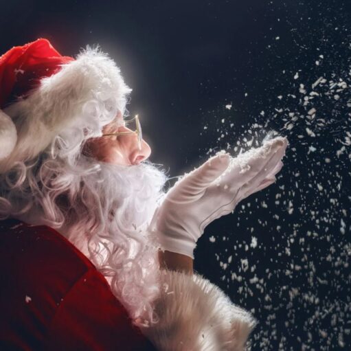 A person dressed as Santa Claus is shown in profile against a dark background, blowing a handful of white flakes into the air. The flakes look like snow or glitter and are sparkling in the light. Santa is wearing his classic red and white suit, a red hat with a white pompom, a white beard, and white gloves. He is also wearing small, round glasses.