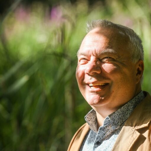 A close-up outdoor portrait of a smiling, fair-skinned man with short grey hair, looking slightly up and to the left. He is wearing a light brown jacket over a patterned collared shirt. Bright sunlight illuminates the right side of his face. The background is a soft, out-of-focus blend of bright green and hints of purple and yellow foliage.