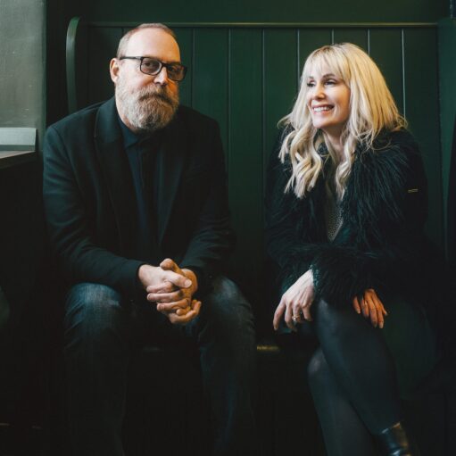 An image of two people, Heidi Talbot and Boo Hewerdine, sitting on a dark green bench indoors. Boo Hewerdine, on the left, has a beard and is wearing a black blazer and glasses. Heidi Talbot, on the right, has blonde hair and is wearing a black faux-fur jacket. Both are smiling.