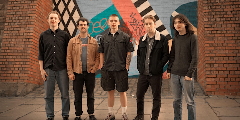 A full-body group portrait of five young men standing in a line in front of a brick wall and a colorful mural. From left to right: a man with a slight smile wearing a black button-down shirt and jeans; a man with a mustache and curly hair wearing a tan jacket over a t-shirt and dark pants with his hands in his pockets; a man in the center wearing a dark gray short-sleeved button-down shirt and shorts; a man wearing a black denim jacket with a white fleece collar and jeans; and a man with long, dark hair wearing a gray sweater and jeans. They all look straight at the camera.