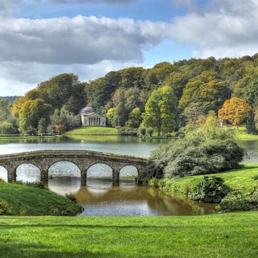 A picturesque landscape featuring a calm lake surrounded by lush, green hills and trees with hints of autumn foliage. A stone arch bridge crosses a narrow part of the water in the foreground, and a small, classical-style temple with a dome is visible on the far bank. The sky is partly cloudy.