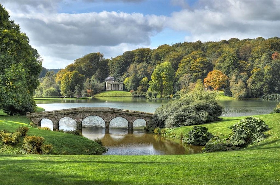 A picturesque landscape featuring a calm lake surrounded by lush, green hills and trees with hints of autumn foliage. A stone arch bridge crosses a narrow part of the water in the foreground, and a small, classical-style temple with a dome is visible on the far bank. The sky is partly cloudy.