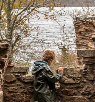 A child in a green jacket and blue hoodie is looking out from behind a stone wall. The stones are part of a ruined structure. Beyond the wall, a large body of water is visible, with trees on the far shore.