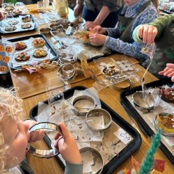 A top-down view of a table where several children are doing a craft activity. They are using cookie cutters and other tools on what appear to be food items or crafts on baking trays. Several children's hands are visible, and the scene is a bit messy, with various supplies and materials scattered across the table.