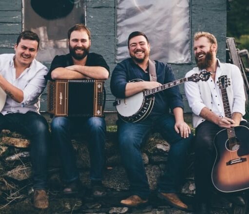 An image of the band Rum Ragged. The four band members are sitting on a stone wall in front of a weathered-looking green building. The man on the left is holding a bodhrán, the second man from the left is holding an accordion, the man to his right is holding a banjo, and the man on the far right is holding a guitar. All four are smiling at the camera.