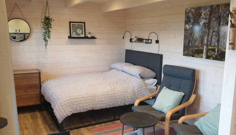 The interior of a cabin bedroom with wood-paneled walls. A queen-sized bed with a gray headboard and a white duvet is centered in the room. A small, round mirror hangs on the wall, and two modern chairs sit next to the bed. A forest landscape picture and a framed print of a plant hang on the walls.