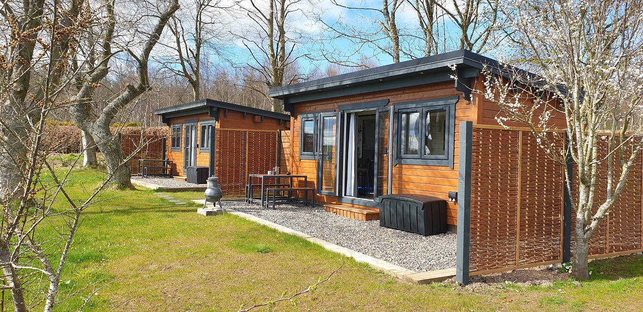Two rustic, wooden cabins with dark gray trim and large windows sit side-by-side in a grassy, outdoor area. Each cabin has a small patio with a table and chairs, surrounded by light-colored gravel. Trees and a small fence are visible in the background under a blue sky.