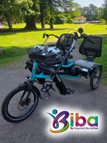 A bright turquoise, three-wheeled tandem bicycle with two seats. The bike has a black storage basket between the two seats and two white helmets are visible in the basket. The front of the bike is in the foreground, facing left. The bike is parked on a paved path in a grassy park, with a building and trees in the background. The logo for BIBA, which stands for "Be Inspired Be Active," is in the bottom right corner of the image.